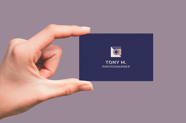A hand holding a deep navy blue business card for 'Tony M. Photographer' with a camera lens logo icon and white typography on a muted purple background. This UV-printed photographer business card uses a premium dark stock with elegant white design — ideal for professional photographers and visual artists seeking a distinctive, sophisticated brand card.