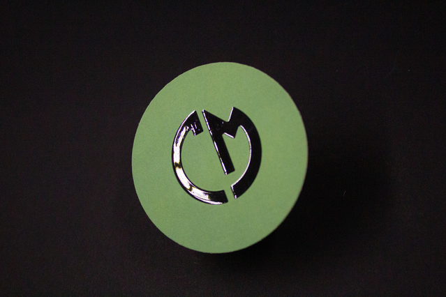 Single round UV business card in vibrant green with a glossy silver spot UV logo at center on a dark background. The bold circular shape and metallic UV embellishment make this card memorable and distinctive. A perfect showcase of premium round UV business card printing.
