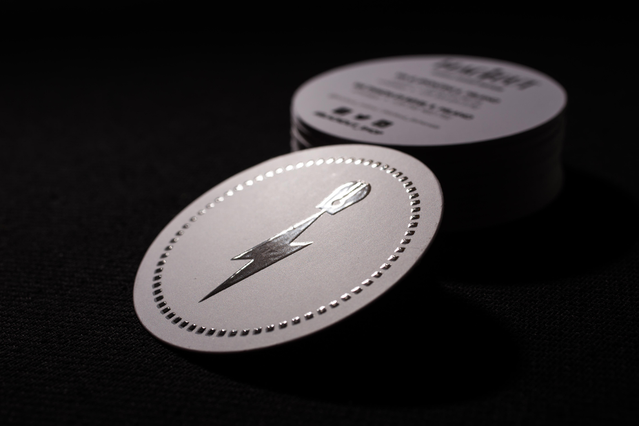 Stack of round UV business cards on dark background, featuring a central silver lightning bolt logo with a dotted border ring on brushed silver stock. The circular die-cut and gleaming spot UV finish create a tactile, high-impact card. A stunning example of round UV business card printing.