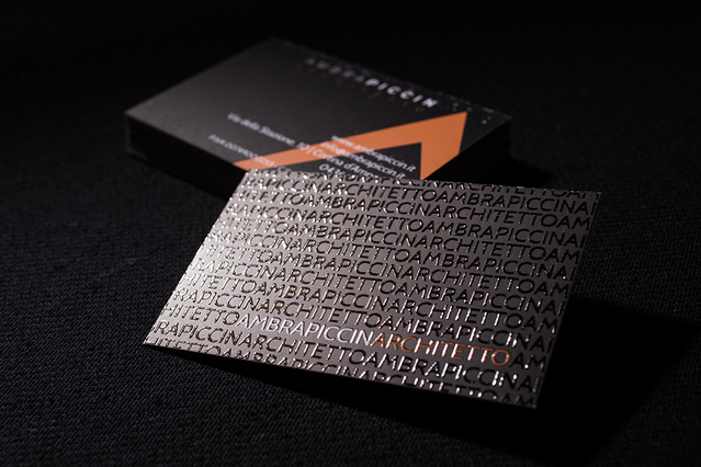 A square dark metallic double-sided business card featuring an all-over repeating embossed text pattern on the front, photographed alongside its black premium packaging box with a copper-orange triangular accent. This double-sided business card print combines special metallic stock with textured relief printing — ideal for luxury brands and creative businesses seeking distinctive double-sided car…